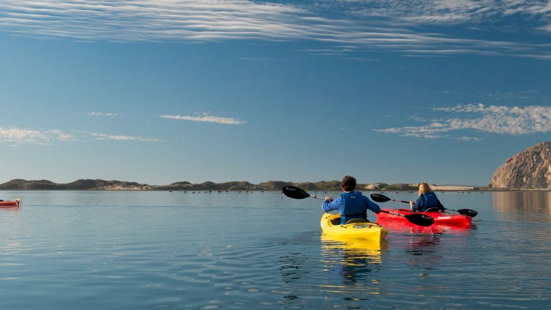 Three people are kayaking on a calm body of water with a large rock formation in the background under a blue sky with scattered clouds.
