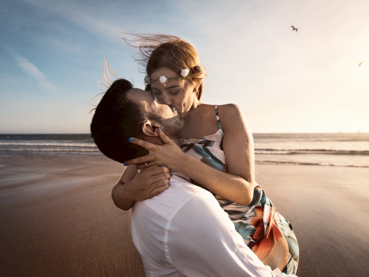 A couple embracing and kissing at the beach with the ocean and birds in the background. The woman wears a floral headband.