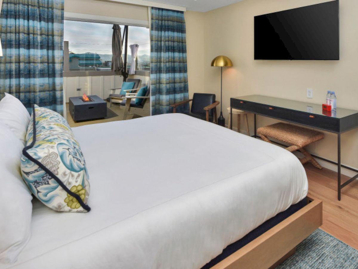 A hotel room with a bed, desk, TV, armchairs, and a large window with drapes. There's a lamp and decorative cushions.