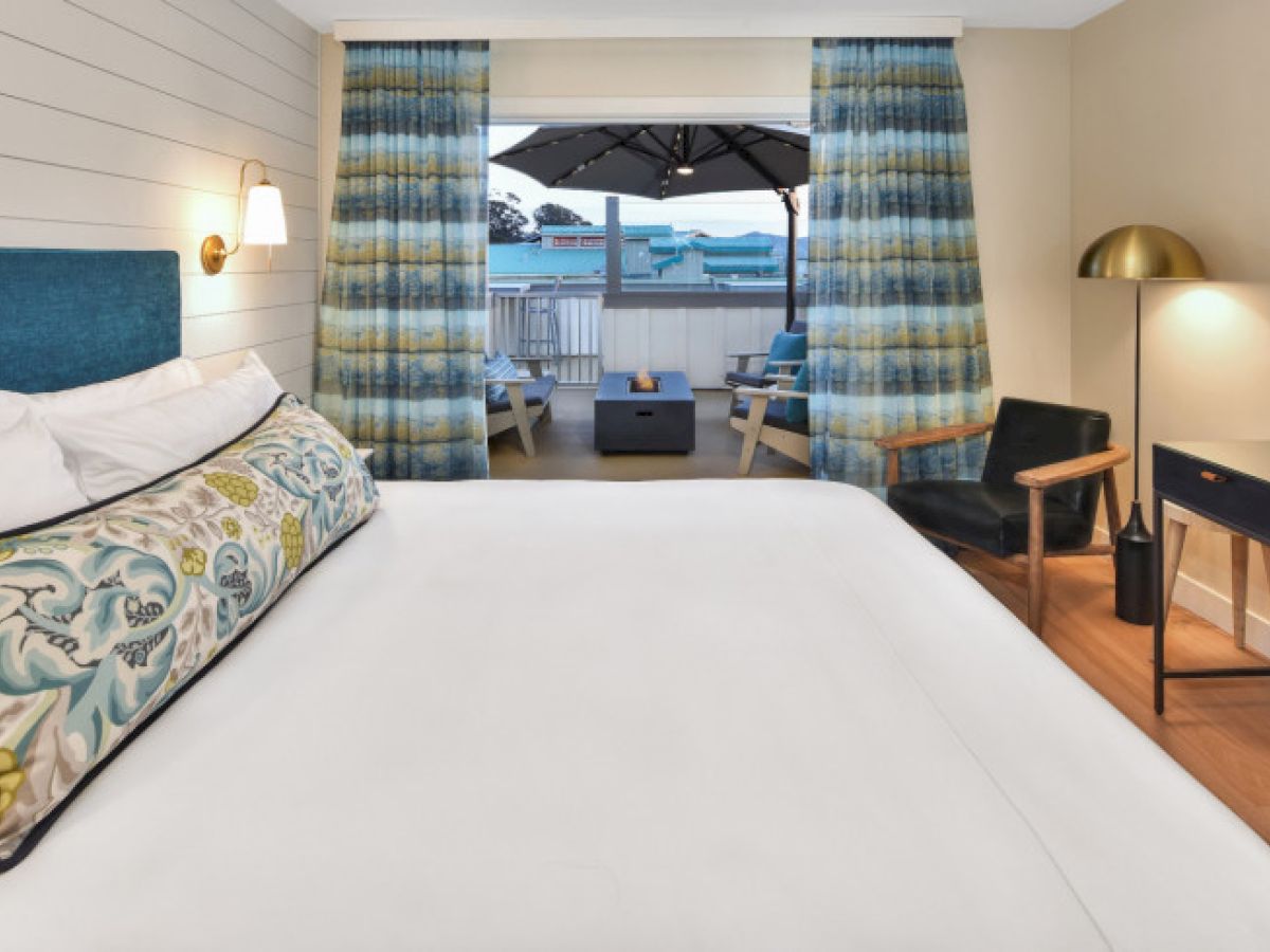 A stylish hotel room features a large bed, desk, chair, and an outdoor balcony with seating and a view, creating a cozy and inviting atmosphere.
