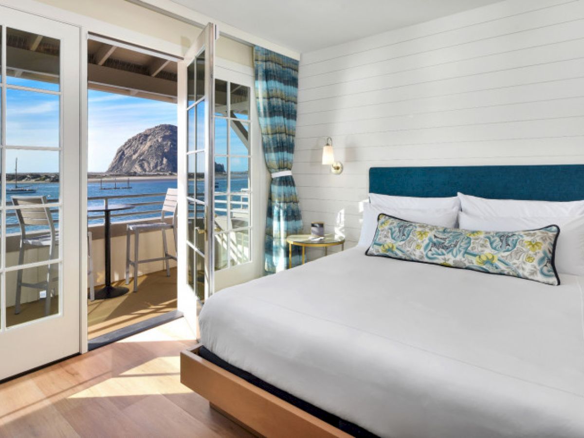 A cozy hotel room features a large bed and an open door leading to a balcony with a stunning ocean view.
