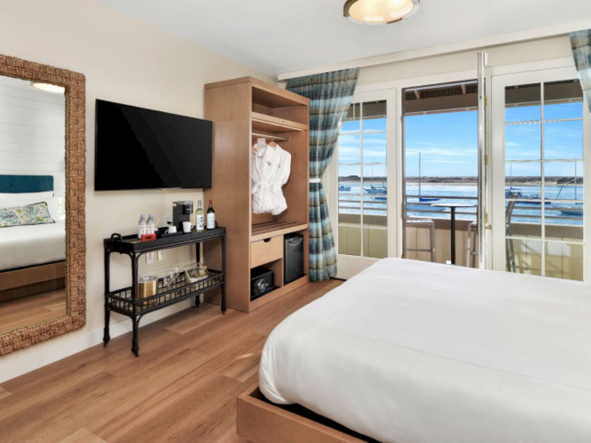 A cozy room with a bed, wall-mounted TV, mirror, and ocean view through large windows and glass doors, with a wardrobe and beverage cart.