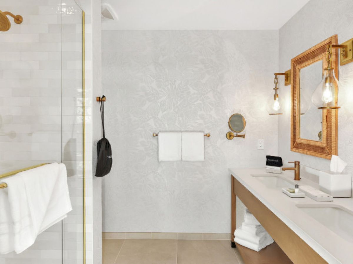 The image shows a modern bathroom with a shower, towels, double mirrors, and a vanity counter with tissue and toiletries on top.