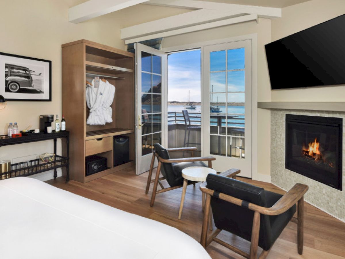 A cozy room has a bed, open closet, TV, fireplace, chairs, and balcony doors with a scenic view of water and a bridge in the distance.