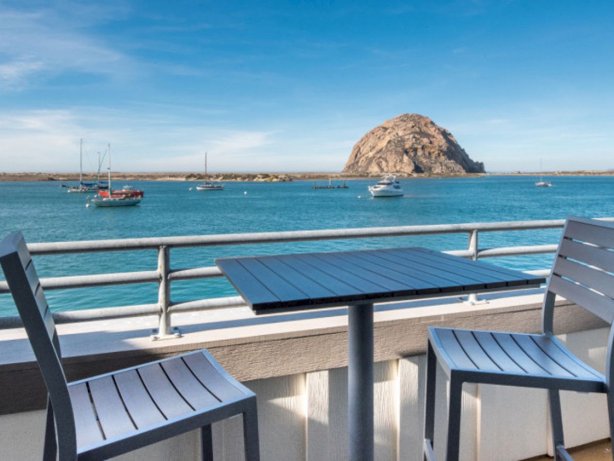 A seaside view with a balcony table and chairs overlooking the ocean, sailboats, and a large rock formation under a clear blue sky.