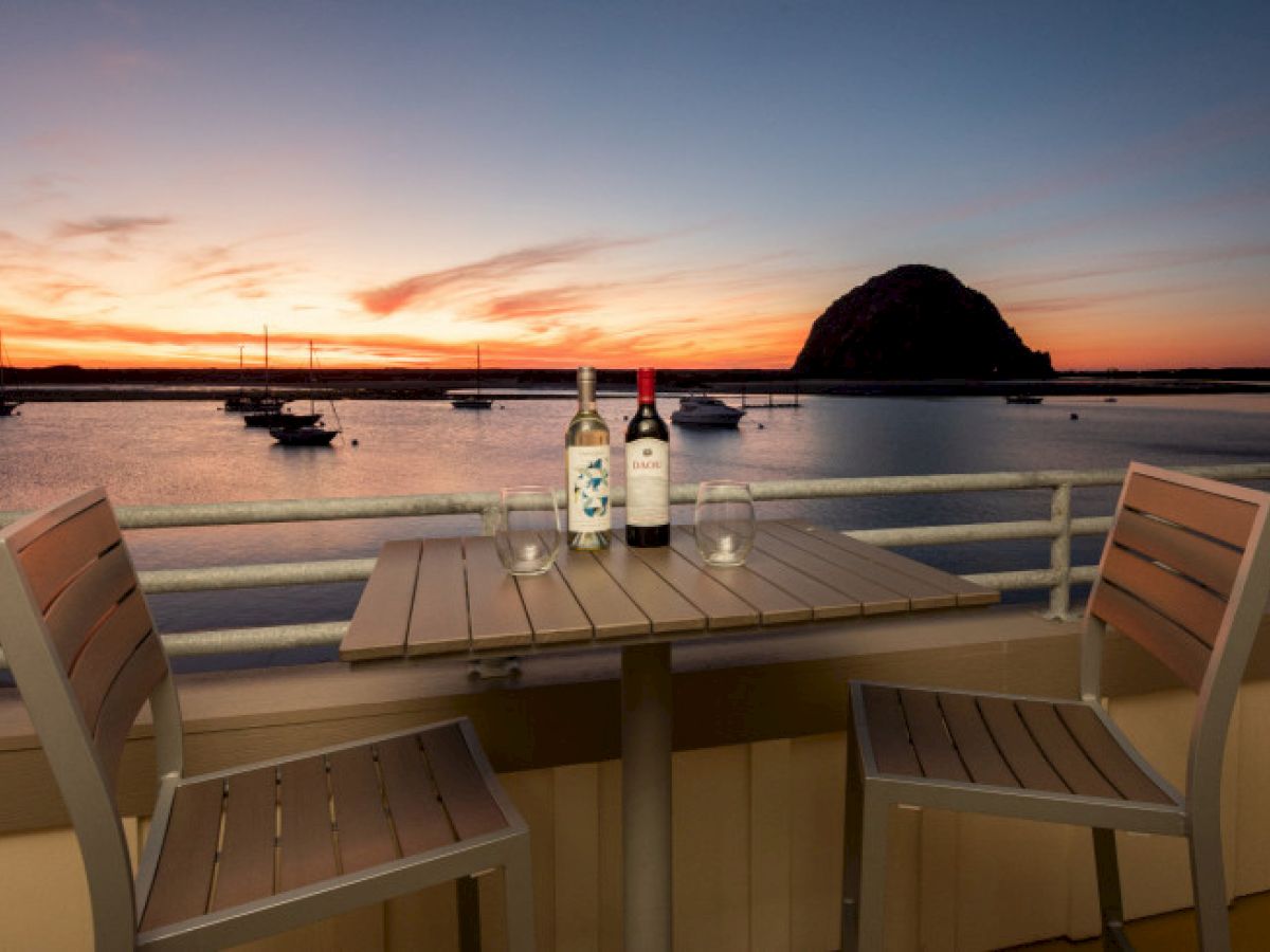 A sunset view over a harbor with boats, featuring a wooden table set with two bottles of wine and chairs on a balcony.