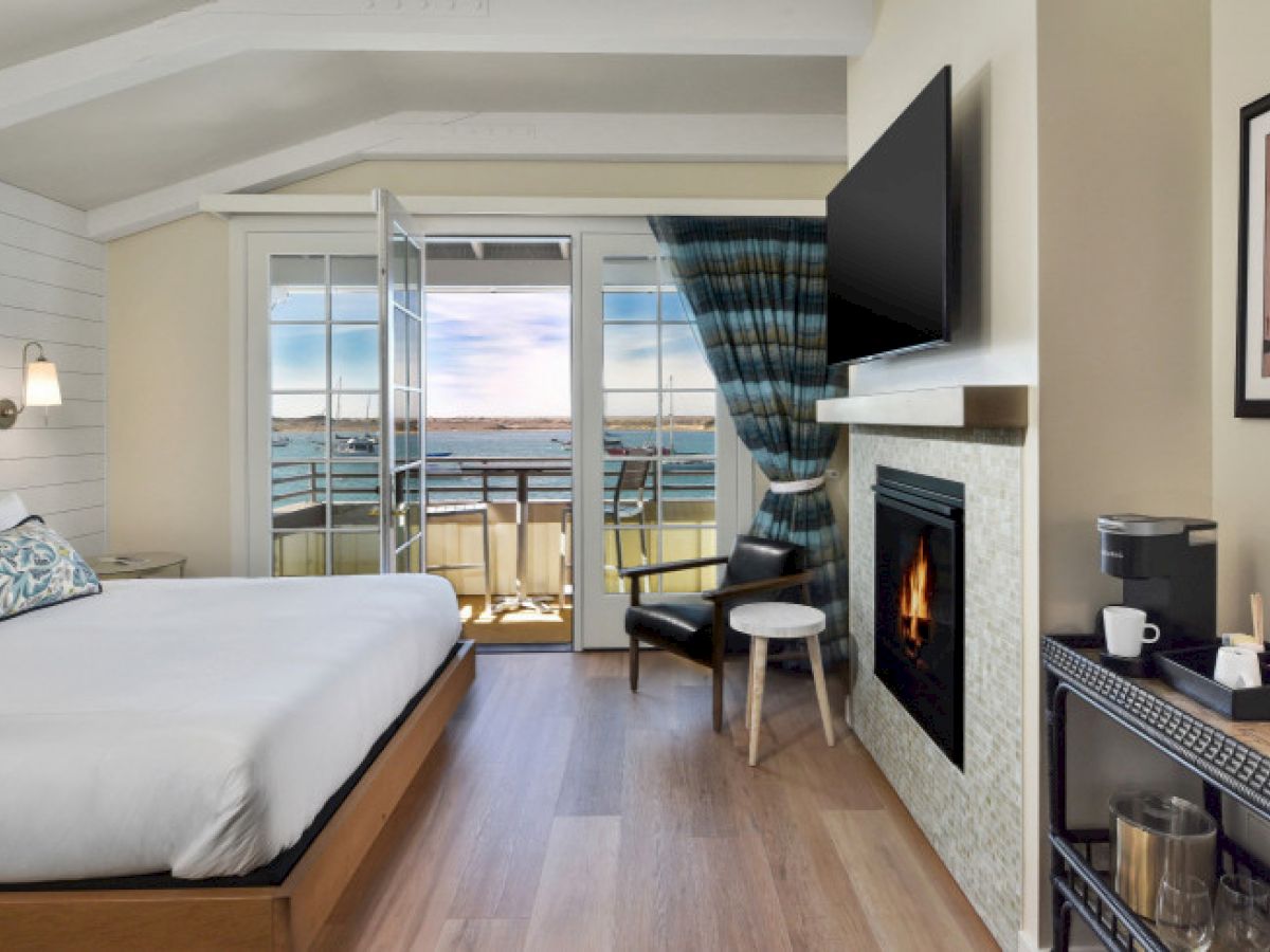 A cozy bedroom features a bed, fireplace, TV, and balcony with ocean view through open doors, creating a serene and inviting atmosphere.