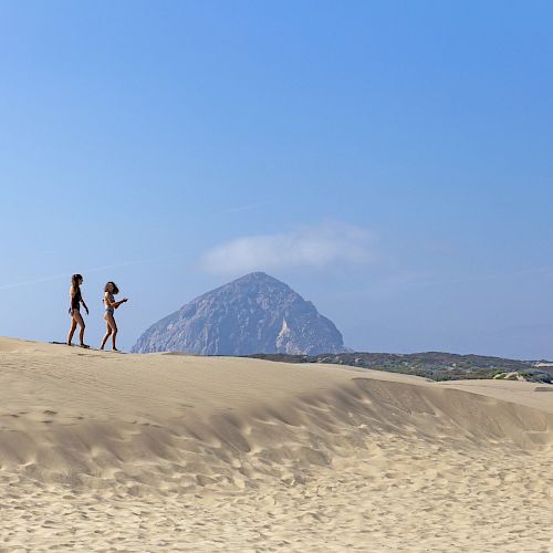 Two people are walking on a sandy beach, with a large rock formation in the distance under a clear blue sky.