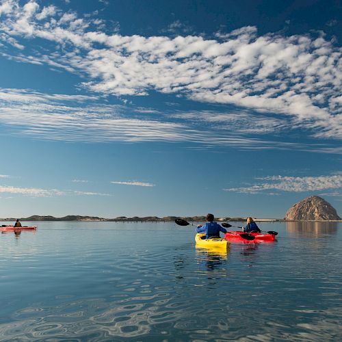 People are kayaking on calm waters under a blue sky with scattered clouds, with a large rock formation in the background.