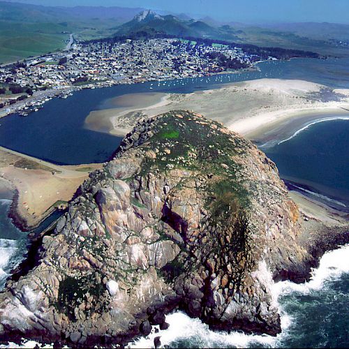 An aerial view showing a coastal town with a prominent rock formation, a beach, a bay, and infrastructure including a power plant.