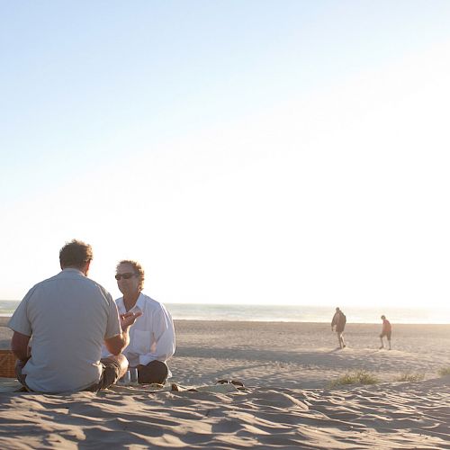 People sitting on sand at a beach during sunset. Two individuals walk by in the background near the water.