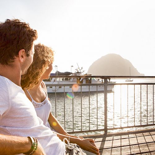 A couple sits on a wooden deck, gazing at a boat on the water with a large rock formation in the background, bathed in warm sunlight.