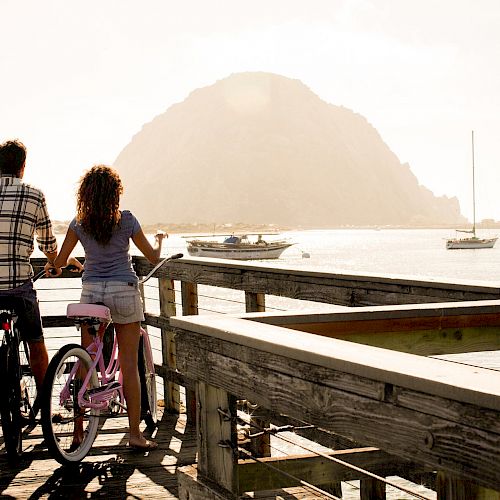 Two people with bicycles stand on a wooden deck overlooking the water with boats and a large rock formation in the background.