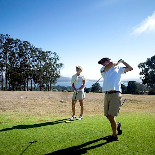 Two people are on a golf course, one is swinging a golf club while the other watches. The landscape includes trees and clear skies ending the sentence.