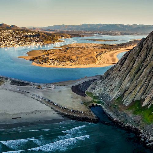 The image shows a coastal landscape with a bay, a town, sandy beaches, and dramatic cliffs bordering the water, all under a clear sky.