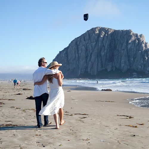 A couple walks on a beach, with the man holding the woman close as she looks at the waves; a large rock formation is in the background.