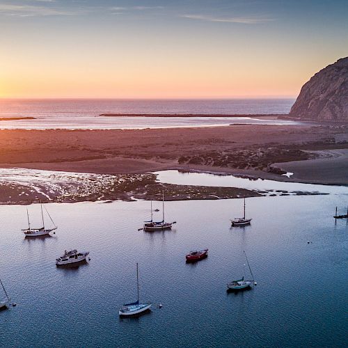 A serene coastal scene at sunset features anchored boats on calm waters, with a magnificent rock formation and coastline in the background.