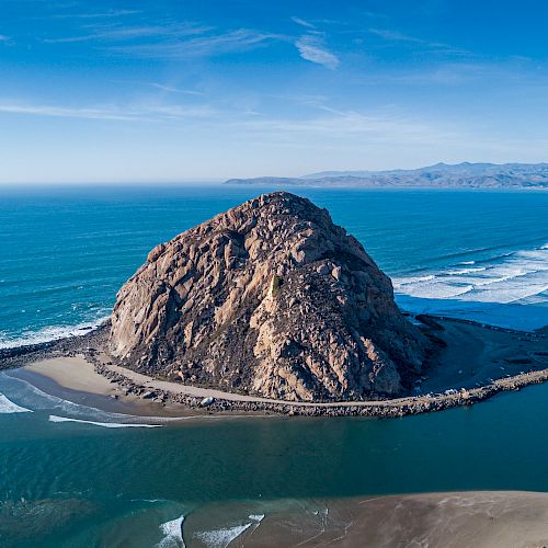 The image shows a large rock formation by the ocean, surrounded by water and sand, with waves crashing on the beach and a mountainous landscape in the background.