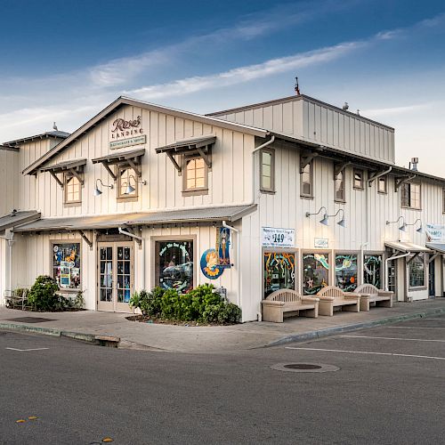 The image shows a white, two-story commercial building with various shops and colorful signs, situated on a quiet street with empty parking spaces.