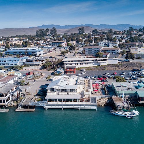 A coastal town with buildings near the water, a docked boat, and hills in the background under a clear blue sky.