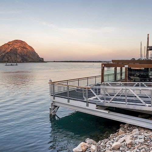 A coastal scene featuring a dockside building, calm water, a visible rock formation in the distance, and a beautifully colored sky.