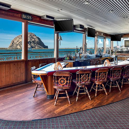 A bar with nautical-themed decor, wooden chairs, and a curved counter overlooking the ocean and a large rock formation through wide windows.