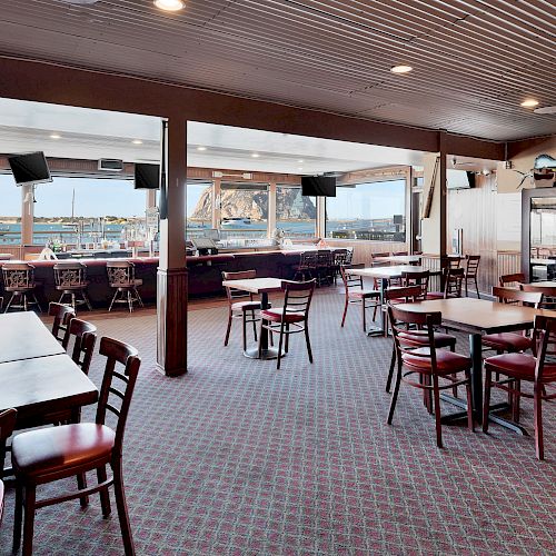 The image shows an empty restaurant or café with tables and chairs, large windows displaying a harbor view, and a bar counter on the left end of the sentence.