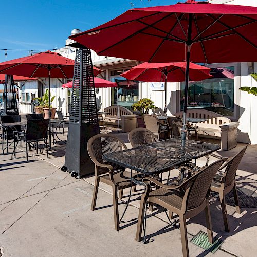 An outdoor seating area with red umbrellas, table and chairs, heaters, and potted plants is shown on a sunny day, next to a white building.