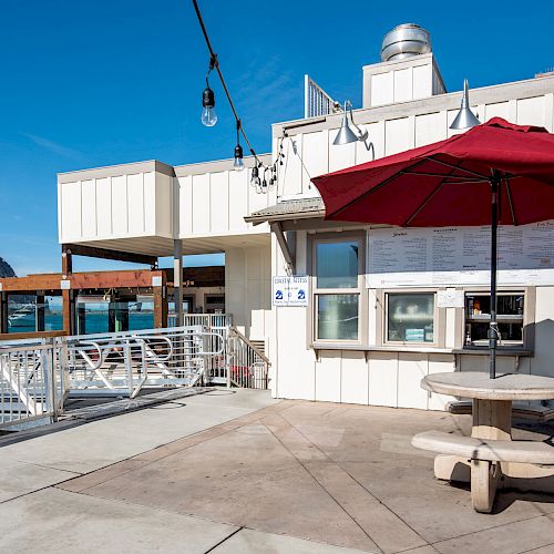 The image shows an outdoor dining area with a round table and a red umbrella beside a white building by the water, with a rock formation in the background.