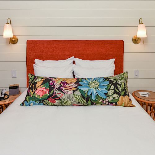 A neatly made bed with a red headboard, floral cushion, two bedside tables, lamps on each side, white pillows, and a shiplap wall background.