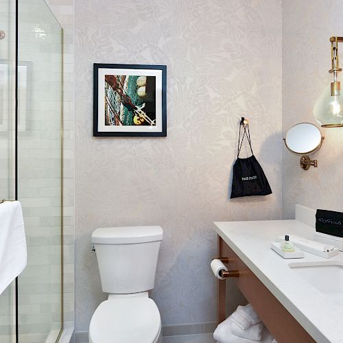 A bathroom with a shower, toilet, sink, and wall mirror. It has a modern design, artwork on the wall, and a towel hanging on the shower door.