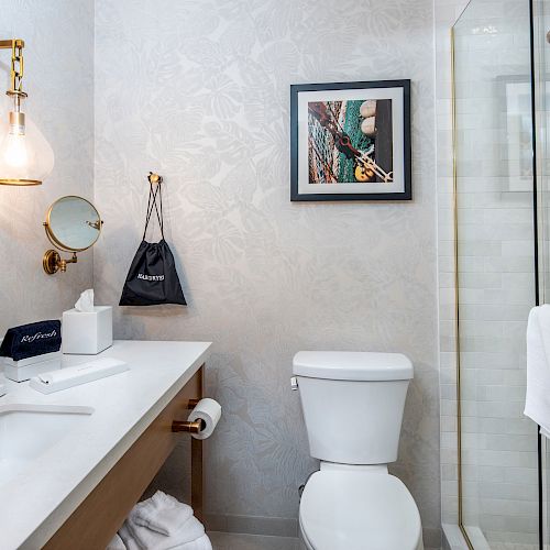 A modern bathroom with a white toilet, countertop, sink, wall mirror, tissue box, hanging artwork, and glass-enclosed shower with a towel.