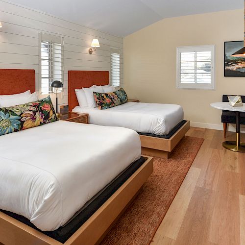 The image shows a hotel room with two white double beds with floral pillows, a small dining table with chairs, wall art, and wooden flooring.