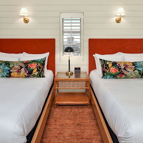 The image shows a bedroom with two beds. Each bed has white bedding, a colorful floral pillow, and a red headboard.