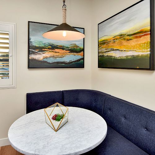 A cozy corner with a round marble table, a blue banquette, pendant light, two landscape paintings, and a window with shutter blinds.