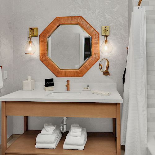 This image shows a modern bathroom with a wooden-framed mirror, sink with faucet, and well-organized towels. Shower with white curtain on the right.