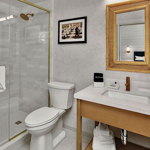 The image shows a modern bathroom with a shower, toilet, sink, large mirror, and a framed picture on the wall. It's tastefully decorated.