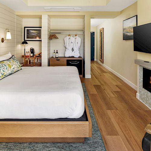 A cozy hotel room features a large bed, a mounted TV, a fireplace, a closet with robes, and a desk with wine and glasses, with modern decor.