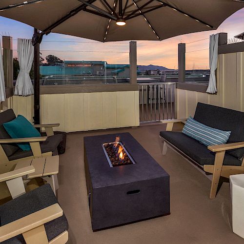 The image shows a cozy outdoor patio setup with cushioned seating, a fire pit, and an umbrella, against the backdrop of a sunset.