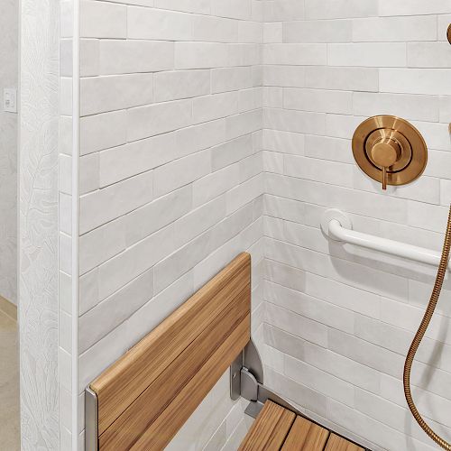 The image shows a shower area with a wooden seat, a grab bar, and a gold-colored shower fixture. Towels are hanging on a rack outside the shower.