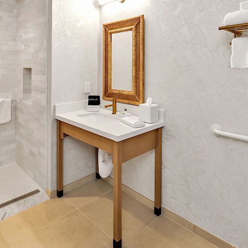 The image shows a clean bathroom with a walk-in shower, a vanity with a large mirror, a sink, and toiletries. A toilet and towel shelves are also visible.