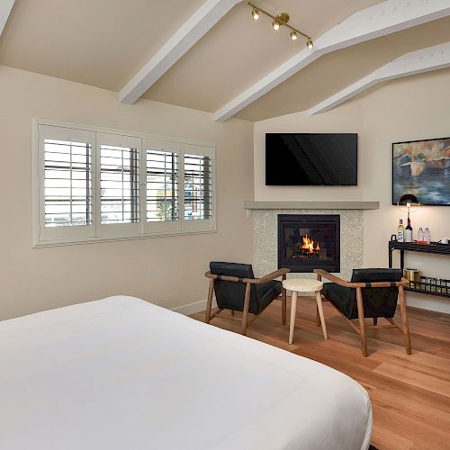 A cozy bedroom features a bed, two chairs, a fireplace, a flat-screen TV, a shelf with amenities, and a wall painting, all under high ceilings.