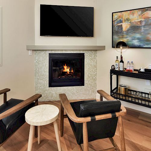 The image shows a cozy corner with two armchairs facing a fireplace, a small table, a wall-mounted TV, abstract painting, and a bar cart.