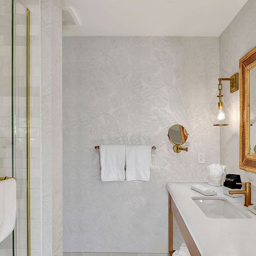 A modern bathroom with a glass shower, a vanity with a gold-framed mirror, a wall-mounted magnifying mirror, and light fixtures.