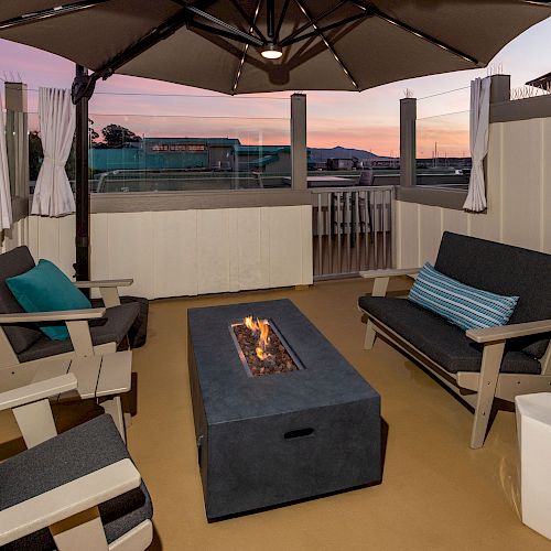 An outdoor seating area with cushioned armchairs, a fire pit table, and a patio umbrella, set against a sunset sky.