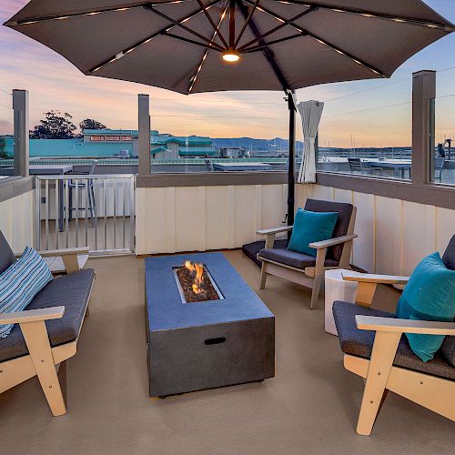 A cozy outdoor seating area with a fire pit table, four cushioned chairs, and an overhead umbrella, set against a sunset sky.