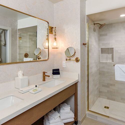 A modern bathroom with a large mirror, dual sinks, a walk-in shower with glass doors, and wooden cabinets below the sink, neatly organized.