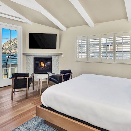 A cozy bedroom with a fireplace, TV, two chairs, and a bed. French doors open to a balcony with an ocean view.