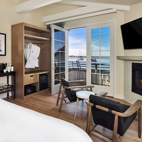 A cozy room features a fireplace, TV, open wardrobe with robes, desk, chairs, and a balcony with a scenic harbor view under a clear sky.