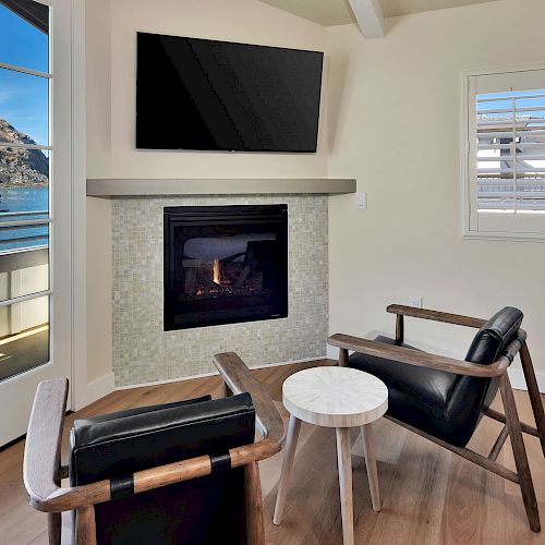 A cozy living room with a wall-mounted TV, fireplace, two chairs, a small round table, and windows displaying a scenic ocean view with mountains.